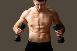 A man is standing and holding two dumbbells in his hands, showing strength and fitness. He has a focused expression and appears to be in a gym or workout space. The mans muscles are visibly tense photo