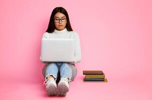 Asian young woman sitting on a pink background while using a laptop. She is looking at the laptop screen and typing. There are three books stacked next to her, copy space photo