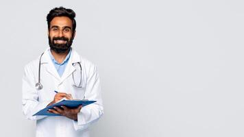 Eastern man wearing a white lab coat and a stethoscope smiles while writing on a blue clipboard with a pen. The background is a plain, light gray, copy space photo