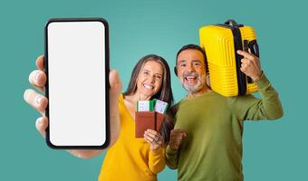 Mature couple stand in front of a green background. The woman holds smartphone with blank screen, passport and tickets, while the man holds a yellow suitcase photo
