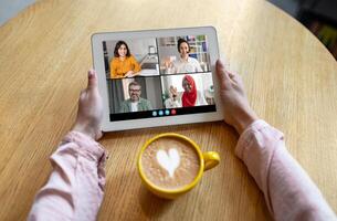 Woman sitting at a table with a cup of coffee and using a tablet for a conference. The tablet screen displays four participants in different squares. photo