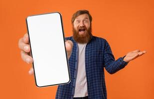 A joyful man with a beard enthusiastically holds up a smartphone with a blank screen, gesturing excitedly against a bright orange background, inviting interaction with a smile. photo