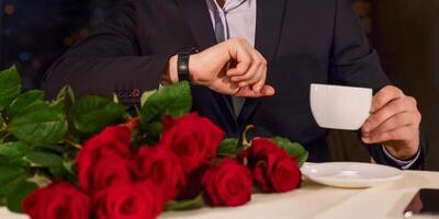 Man with flowers waiting girlfriend in restaurant nervous, drinking coffee, checking time. She is late concept photo