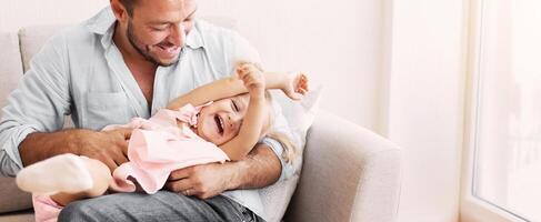 Best Dad In The World. Millennial man tickling his daughter who lying on his lap on the sofa. Free space photo