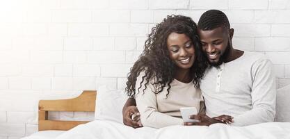Smiling black couple using smartphone sitting on bed together, checking social networks or browsing media, copy space photo