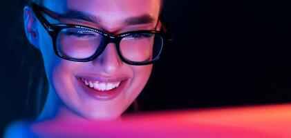 A woman wearing glasses is sitting in front of a computer screen, smiling brightly as she looks at the display. Her expression conveys happiness and engagement with the content on the screen. photo