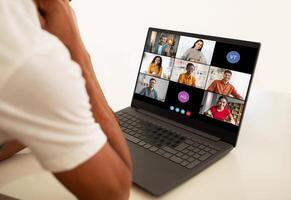A man sits at a desk, looking intently at his laptop screen. He is participating in a conference with a diverse group of people. The screen displays multiple feeds photo