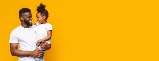 Black father is holding his young daughter in his arms, both beaming with happiness. They are positioned in front of a bright yellow backdrop, creating a cheerful and lively atmosphere, copy space photo