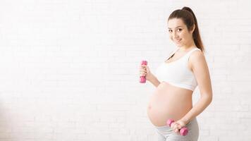 A pregnant woman is standing while holding two pink dumbbells in each hand. She appears to be focused and engaged in her workout routine, maintaining balance and stability, copy space photo