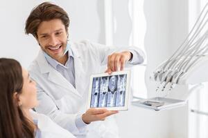 Modern technologies in dentistry. Handsome cheerful dentist doctor showing his patient x-ray results on digital tablet, copy space photo