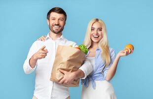 A man and a woman stand together against a blue background. The man is holding a brown paper bag filled with groceries and pointing to the side with his right hand photo