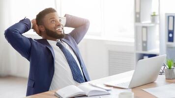A happy businessman in a suit sits at his desk with his hands behind his head, leaning back in his chair with a laptop in front of him. photo