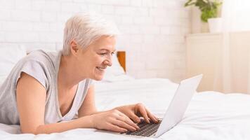 Senior woman is laying on a bed, fully engrossed in using a laptop computer. She is positioned comfortably with the laptop in front of her, typing and browsing, copy space photo