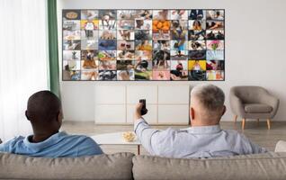 Two men, one black and one white, are sitting on a couch in a living room, facing a large television screen. The screen is showing a grid of images, creating a mosaic effect. photo