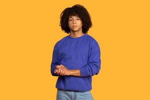 A young man with curly black hair is wearing a blue sweatshirt and standing in front of a yellow background. He is looking directly at the camera with a neutral expression. photo