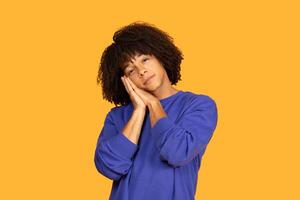 A young man with curly hair is wearing a blue sweater and posing as if he is sleeping, with hands together under his cheek. He stands in front of a vibrant orange background photo