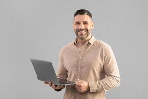 A man is holding a laptop computer in his hands, looking at the screen with a smile on his face. He appears engaged and content, possibly working or communicating with someone through the device. photo