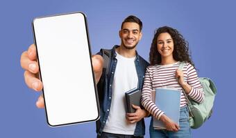 Two arabic students are standing in front of a blue background. They are both smiling and holding books and backpacks. The man is holding a smartphone with a blank screen in front of him. photo