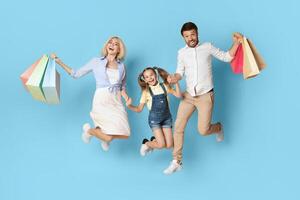 A happy family of three, a mother, father, and young daughter, all wearing casual clothes and holding shopping bags, jump in the air in front of a blue background. photo