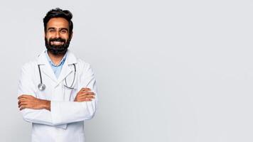 Indian man doctor with a dark beard and a white lab coat stands against a white background with his arms crossed and smiling at the camera. He is wearing a stethoscope around his neck, copy space photo