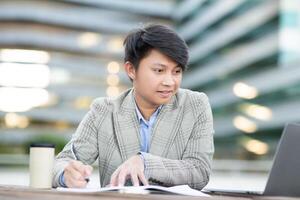 A young Asian man sits at a table outside an office building, working on his laptop. He is writing in a notebook with a pen, and his laptop is open in front of him. photo