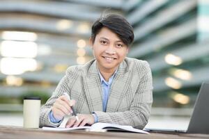 Asian man wearing a plaid jacket is sitting outside a modern building. He is writing in a notebook and looking directly at the camera with a smile. A laptop and coffee cup are on the table photo