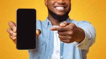 Check this app. Cheerful afro guy holding and pointing at smartphone with black screen, yellow background with free space photo