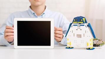 Boy sits at a table and holds up a tablet with a blank screen. To the right of the tablet sits a small, blue and white robot toy with wheels. photo