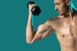 A man is standing in a gym setting, holding a pair of dumbbells and posing for the camera. The background is a solid blue color, emphasizing the man and his workout equipment. photo