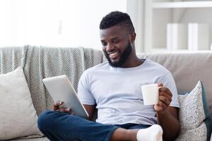 Black man sits comfortably on a couch, his legs crossed, holding a digital tablet in one hand and a mug of coffee in the other. He smiles as he appears to be engaged in something on the tablet. photo