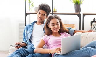 Black couple relaxes on a couch in their home. The girl is using a laptop computer while the guy watches television with a remote control in his hand. They both appear happy and content. photo