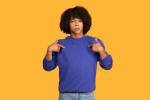 A young man wearing a blue pullover is seen pointing towards himself while standing against a vibrant yellow background. His facial expression suggests he is emphasizing or making a point. photo