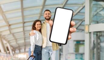 A cheerful man and woman stand together in an airport terminal, with the man extending his arm towards the camera, showing a smartphone with a blank screen photo