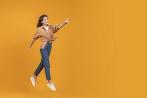 This image shows a woman in a brown shirt and blue jeans jumping in the air while pointing her right hand to the left side of the frame, against a yellow background, copy space photo