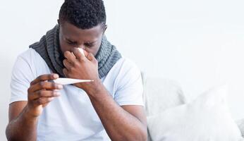 A sick man sits on a couch, wearing a gray scarf, holding a thermometer and tissue. He looks down at the thermometer, and his hand is on his nose as if he is blowing it. He looks upset and sad. photo