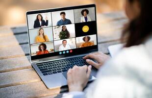 Unrecognizable woman sitting at a desk, typing on a laptop while conferencing with a diverse group of people displayed on the screen. The individuals on screen are engaged in a virtual meeting photo