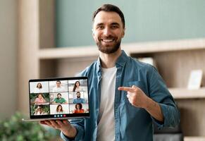 A man is holding a laptop displaying a group of people. He is engaging with the content on the screen while the individuals on the tablet appear focused and engaged as well. photo