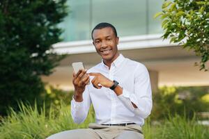A young black man, wearing a white long-sleeved shirt and khaki pants, sits outdoors in front of a modern building with lush greenery surrounding him. He is smiling and using his smartphone photo