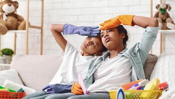 Tired black sisters with cleaning gloves on sitting on sofa, having break while cleaning, empty space photo