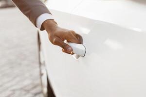 A close-up image showcasing a mans hand opening the door of a white car. The hand is reaching for the door handle, which is visible in the center of the frame. photo