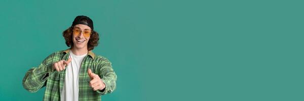 Portrait of cool young guy in sunglasses pointing at you on turquoise studio background, panorama with copy space photo