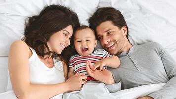 This image shows a happy family of three in bed together during the day. The parents are smiling and looking at their baby, who is laughing and reaching out to touch his fathers face. photo