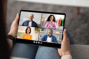Woman is holding a tablet in her lap, participating in a chat with four other people. The call is split into four screens, each showing a different person. photo