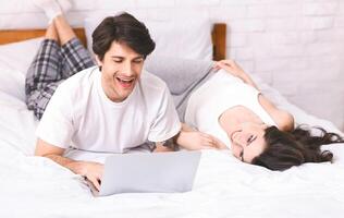A man and a woman are relaxing in bed together. The man is using a laptop and smiling while looking at the screen. The woman is lying on her back with her head turned to look at him, smiling. photo
