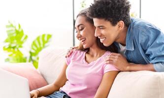 Black couple sits on a white couch in a brightly lit room. The girl is on the left side of the couch, using a laptop while the guy leans in from behind her, smiling and looking over her shoulder photo