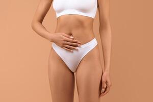 A woman is wearing white underwear and has her hands resting on her stomach. The background is a muted orange color, cropped photo