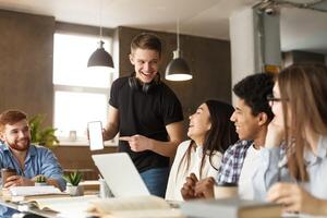 A group of young adults is gathered around a table in a well-lit co-working space, engaging in a lively discussion. One man stands showing his smartphone to the group photo