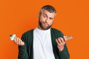 Confused bearded man holding smartphone and credit card in his hands and grimacing, isolated on orange studio background photo