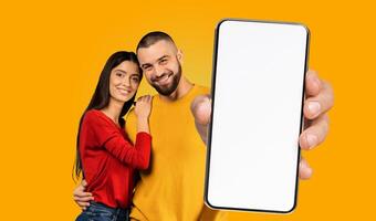 A happy young couple stands together in front of a yellow background, smiling at the camera. The man is holding a smartphone with a blank white screen in front of them. photo
