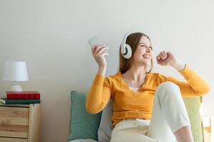 A girl is seated on a bed, wearing headphones and listening to music. She appears relaxed and focused on the music playing. The room is simple, with a bed and a few personal items visible. photo